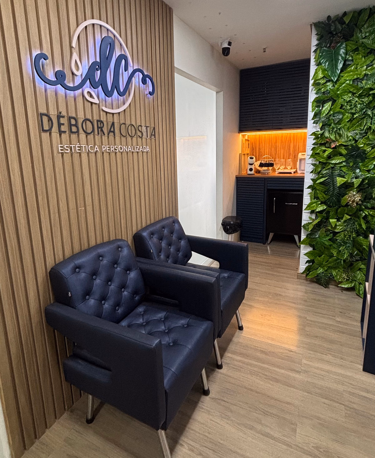 Recepção do studio: letreiro retroiluminado com o monograma e o nome Débora Costa — Estética Personalizada numa parede de ripas de madeira clara, duas poltronas azul-marinho capitonê à frente e uma parede de folhagem à direita.