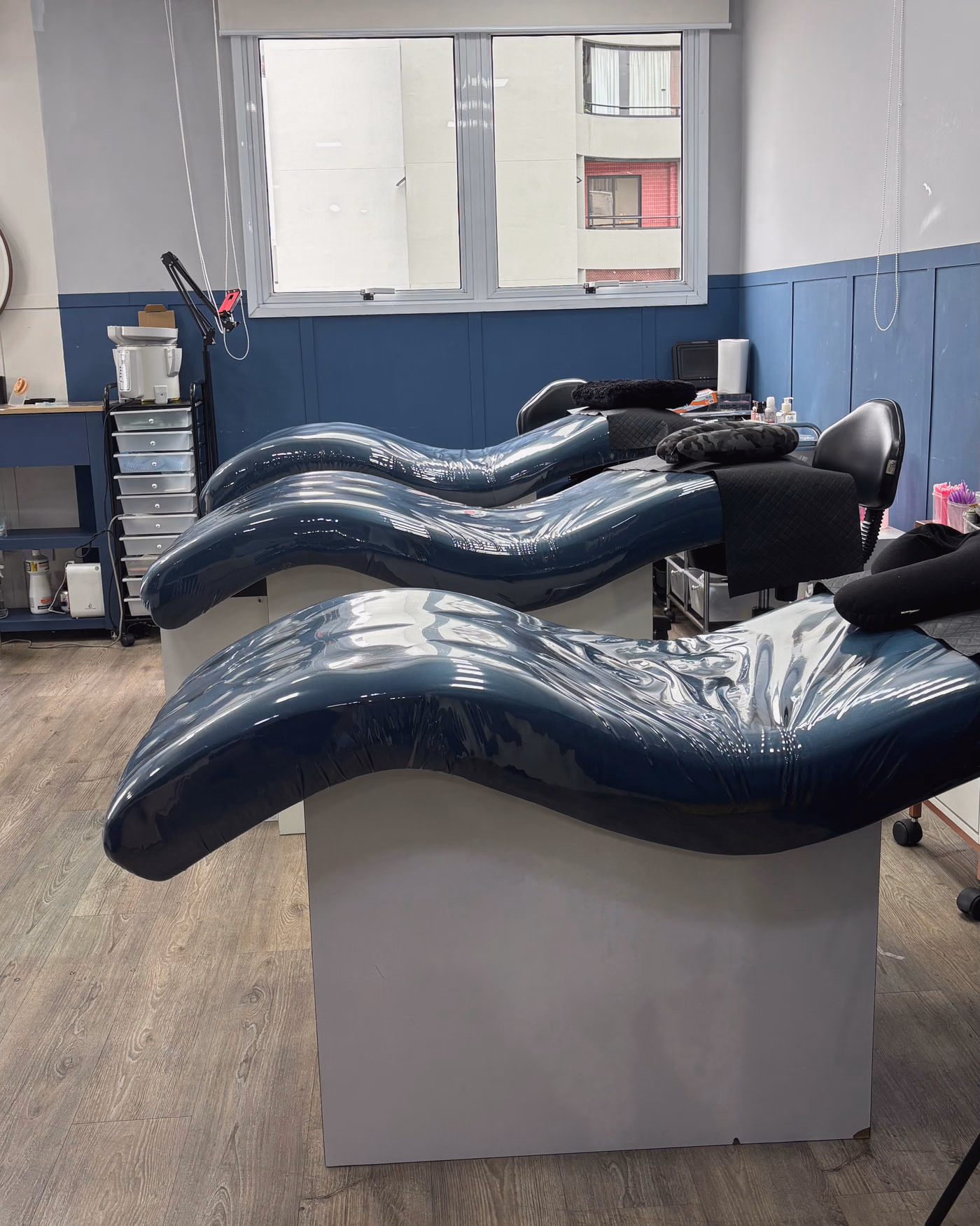 Sala de procedimento do studio: três macas de encosto ondulado revestidas em vinil azul-petróleo, parede com lambri azul-marinho, janela com persiana ao fundo e carrinho auxiliar de gavetas à esquerda.